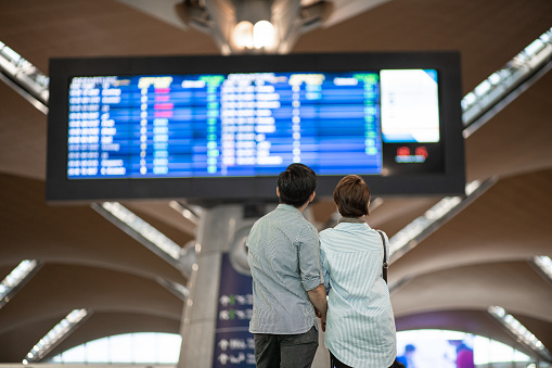 Rear view. Asian Chinese couple checking boarding time in airport. Waiting in front arrival departure board.